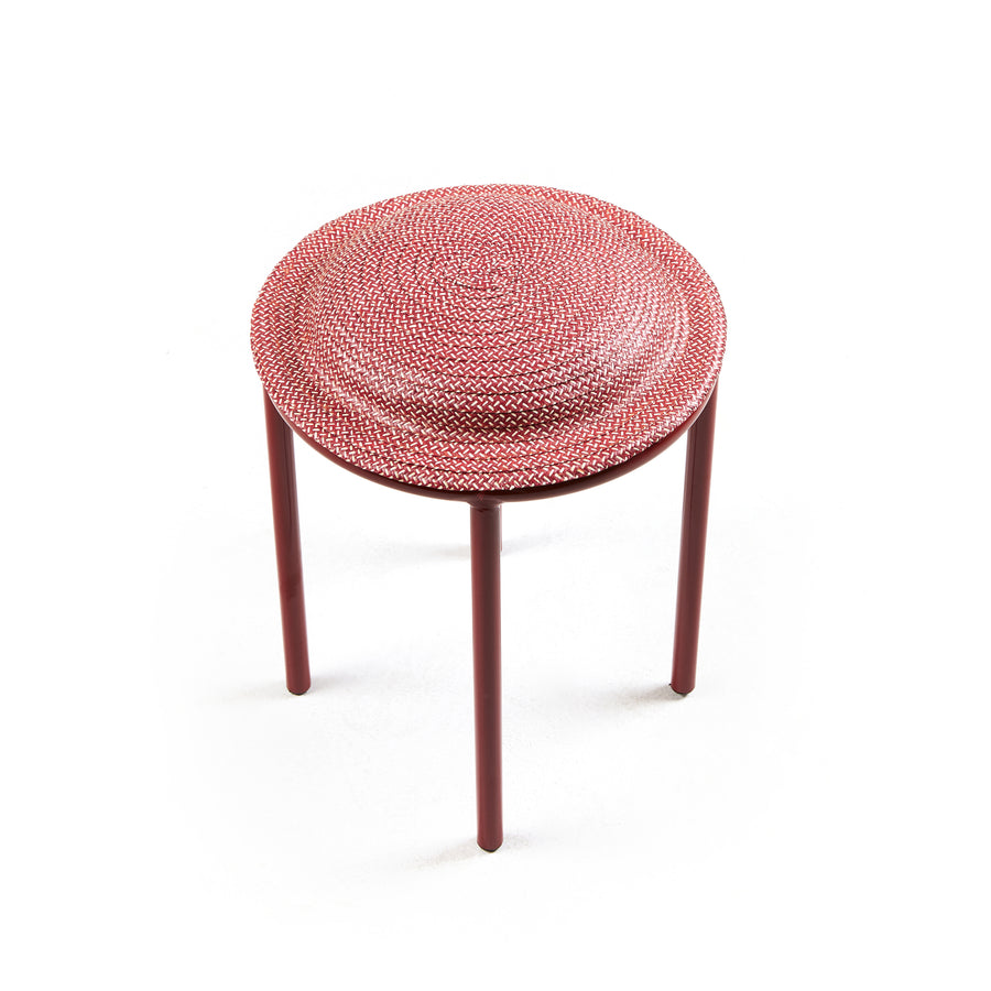 The red Ames Cana Stool boasts a woven circular seat design, contrasting its simple cylindrical legs against a white backdrop. Its vibrant colors and distinctive craftsmanship create an eye-catching, unconventional look.