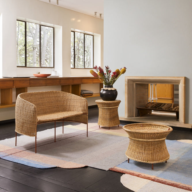 A modern living room with large windows, light walls, and dark wood floors features natural wicker furniture, including the Ames Caribe Natural Basket Table by Ames, a bench, a black vase with flowers, a geometric rug, shelves, and a decorative fireplace.