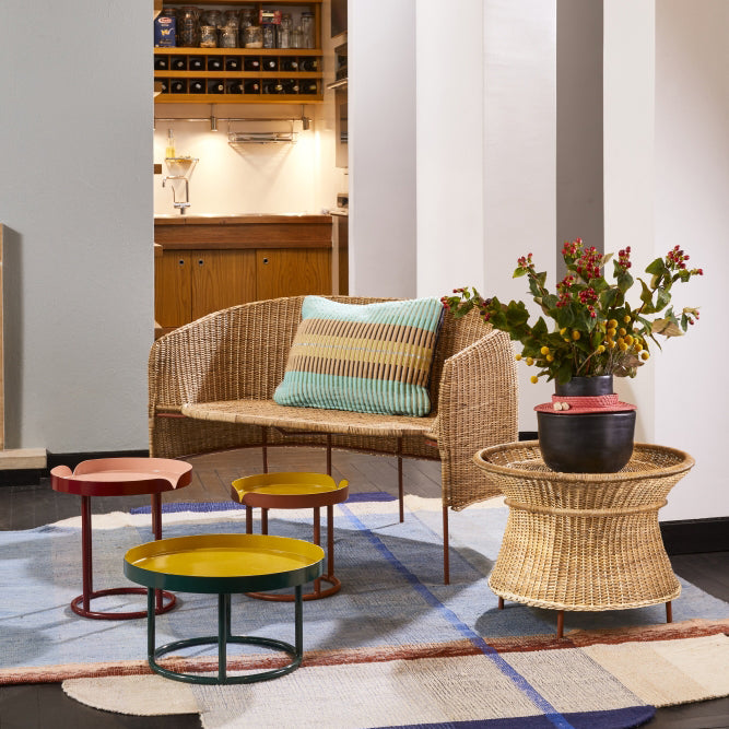 A cozy living space with natural wicker furniture, including a loveseat with a striped cushion, three colorful round side tables, the Ames Caribe Natural Basket Table by Ames holding a potted plant with red berries, and a light patterned rug. A warm kitchen glows in back.
