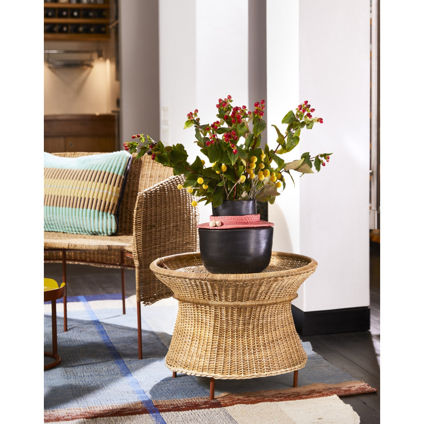 The Ames Caribe Natural Basket Table by Ames sits in a cozy living room with two potted plants, next to a natural wicker chair with a colorful striped cushion and a blue patterned rug beneath.