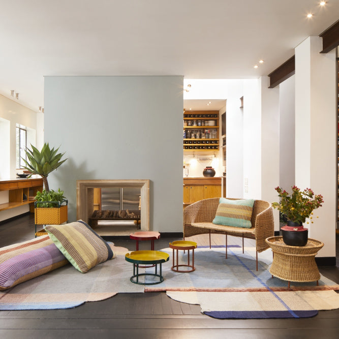 A cozy living room showcases a modern fireplace, vibrant rugs, cushions, and a Caribe Natural Sofa by Ames highlighting Colombian craftsmanship. A potted plant rests on the left side next to a small table, with another plant on the right allowing a view into the kitchen beyond.