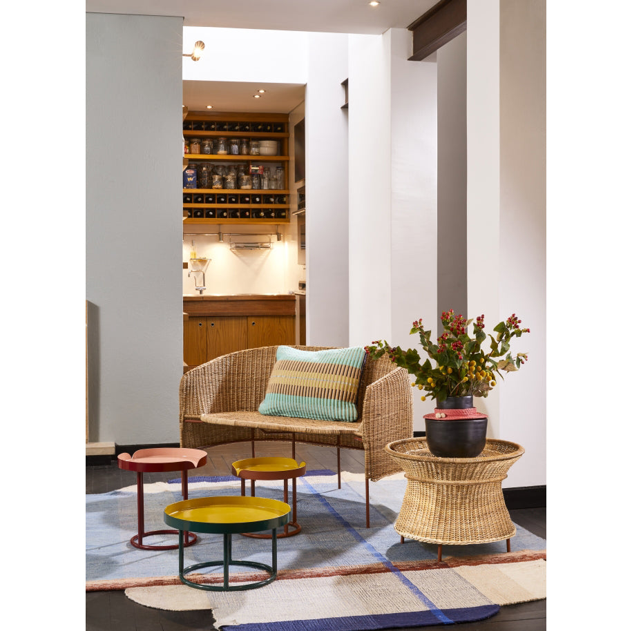 A cozy living area features the Caribe Natural Sofa by Ames adorned with a striped cushion, surrounded by vibrant, colorful round tables. A woven basket holds a red flowering plant. The floor showcases Colombian craftsmanship with a geometric rug, leading to the softly lit kitchen in the background.