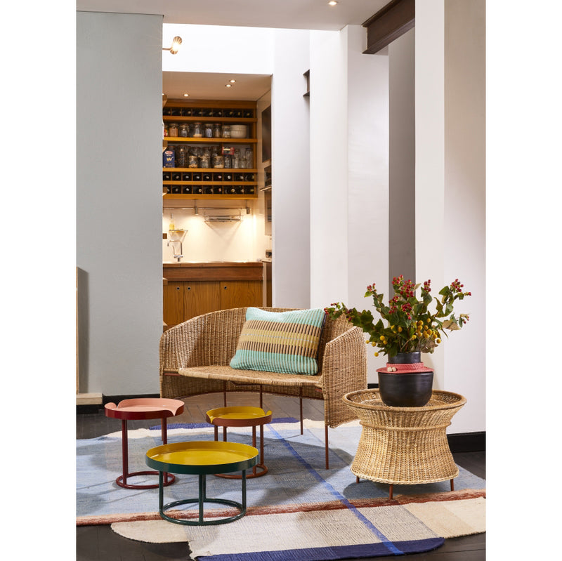 A cozy living area features the Caribe Natural Sofa by Ames adorned with a striped cushion, surrounded by vibrant, colorful round tables. A woven basket holds a red flowering plant. The floor showcases Colombian craftsmanship with a geometric rug, leading to the softly lit kitchen in the background.