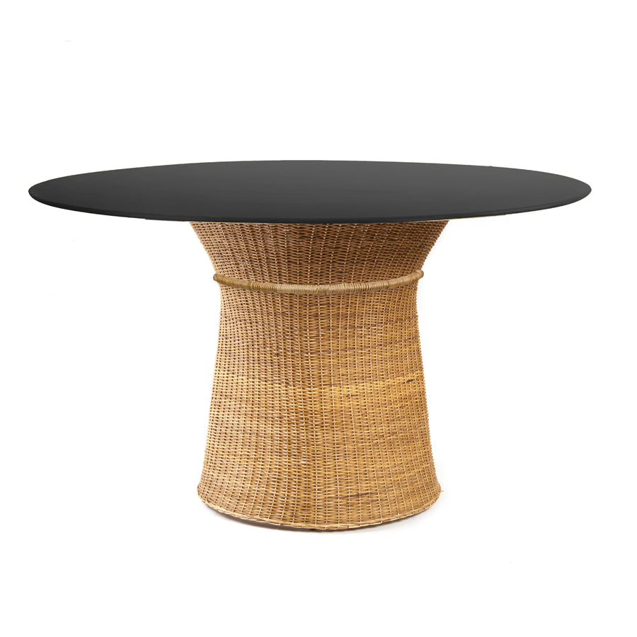 The Ames Caribe Natural Dining Table by Ames features a smooth black round top and a natural woven rattan pedestal base inspired by Sebastian Herkner, shown isolated on a white background.