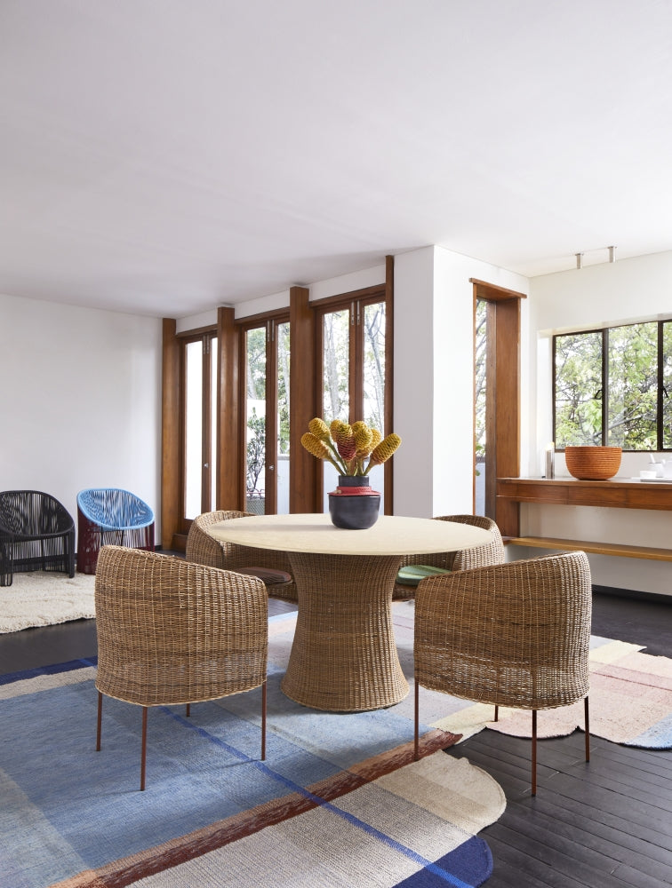 A bright dining area features the Ames Caribe Natural Dining Table by Ames, three matching chairs, a vase with yellow flowers, large windows, neutral-toned decor, and a patterned rug. Natural light fills the modern space.