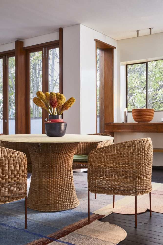An Ames Caribe Natural Dining Table by Ames, paired with matching chairs, sits on a patterned rug. A black vase with yellow and red flowers rests on the table. Large wood-trimmed windows fill the cozy, modern room with light.