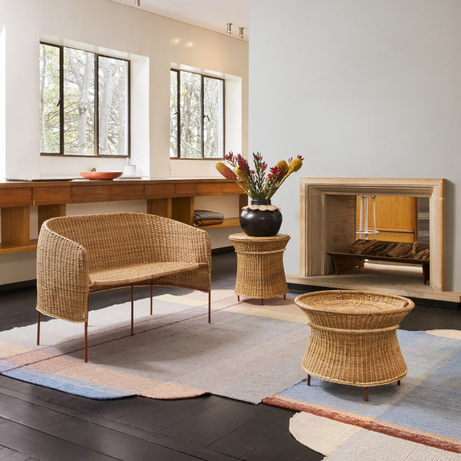 A cozy living room features natural wicker furniture, including a wicker chair and two matching tables, highlighting Colombian craftsmanship. A vase with flowers sits on one. Large windows showcase tree views, and a fireplace enhances the wall, while neutral tones and light add warmth. The high side table is the Caribe Natural by Ames.