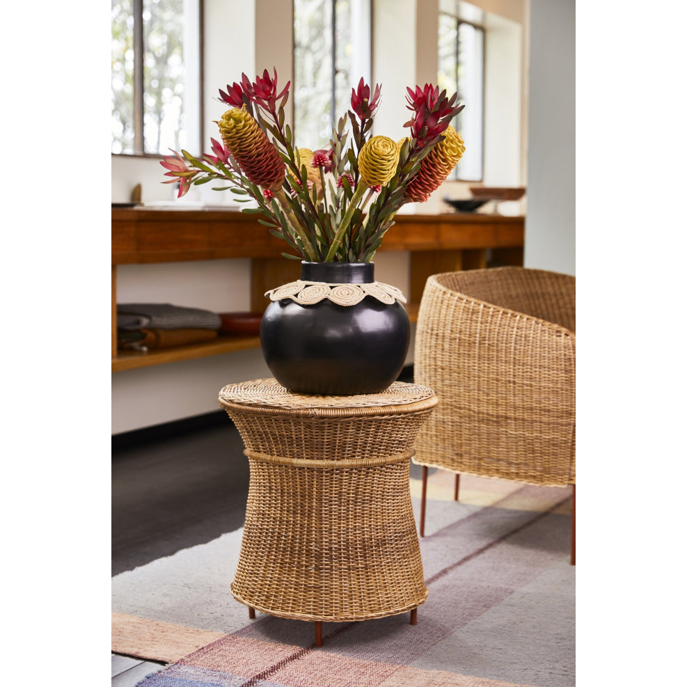 A dark vase with red and yellow tropical flowers sits on the Ames Caribe Natural High Side Table in a bright room. Designed by Sebastian Herkner, the tables natural wicker material highlights Colombian craftsmanship, while a large window allows ample natural light to flood the space.