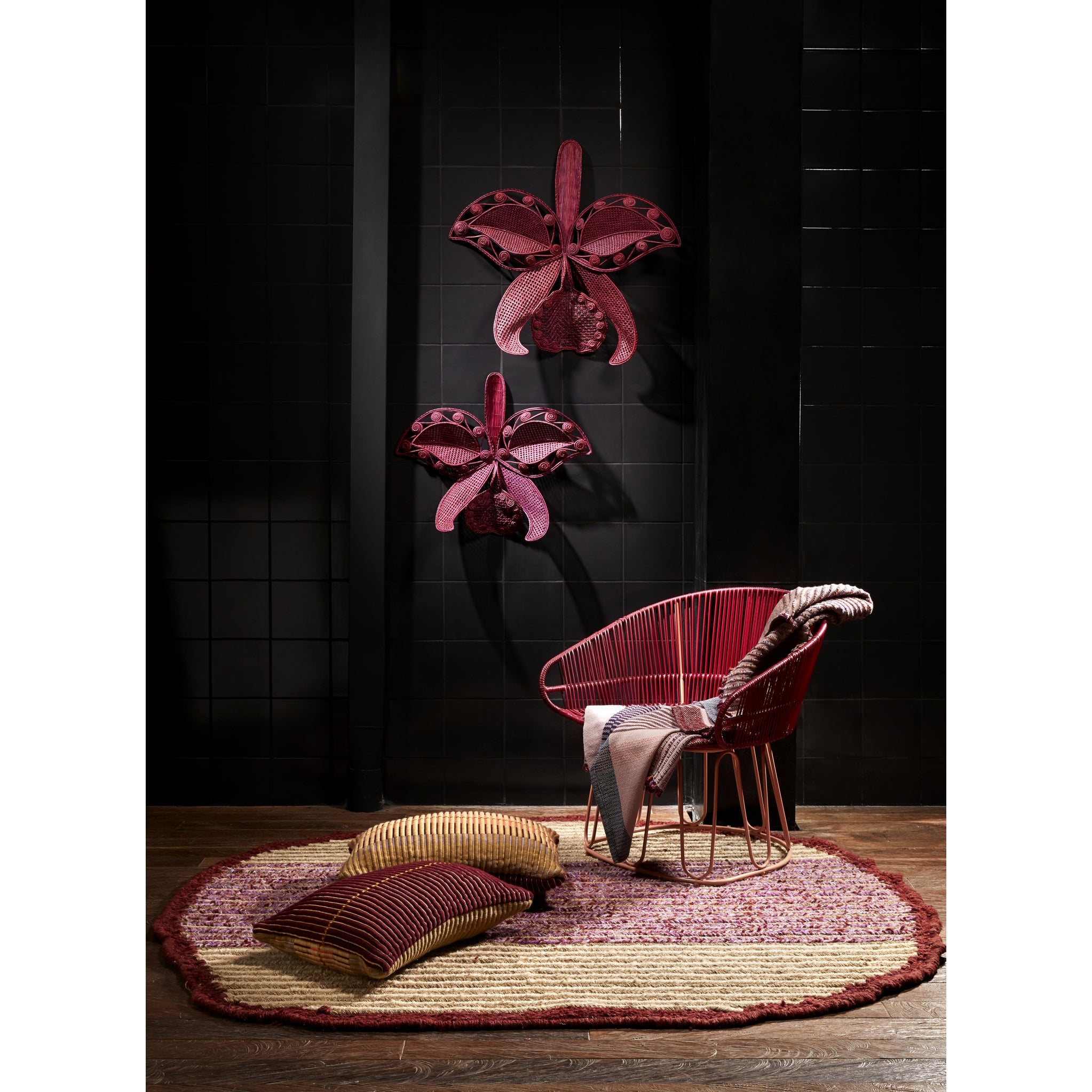 A modern room with black tiled walls, two large Ames Cattleia Wall Decor pieces by Ames, a red wire chair draped with a blanket, two striped pillows, and a round textured rug on a wooden floor.