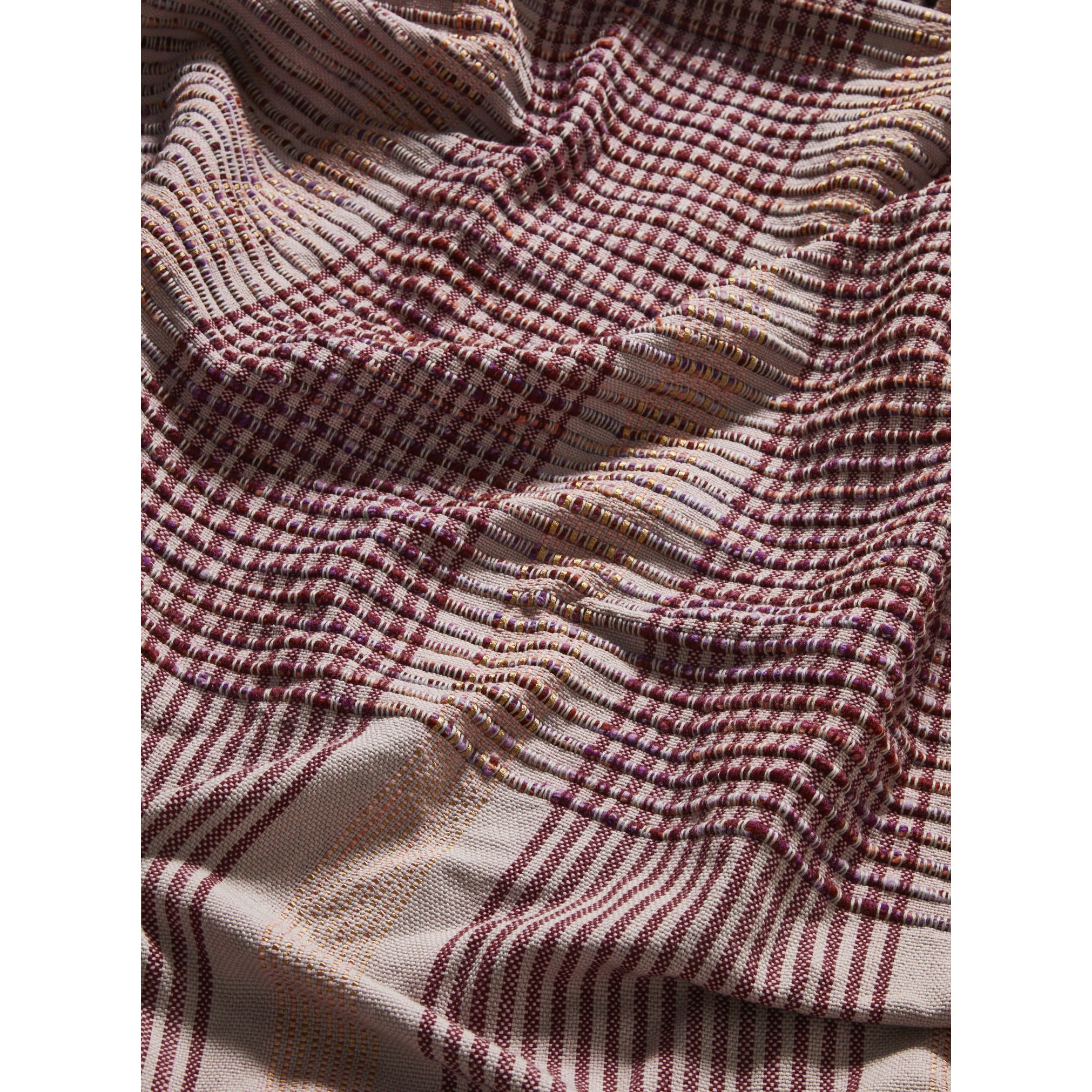 A close-up of a Chumbes Throw by Ames highlights its textured plaid fabric, featuring a red, black, and beige checkered pattern with small stripes. The soft, slightly crumpled material creates intriguing light and shadow effects.