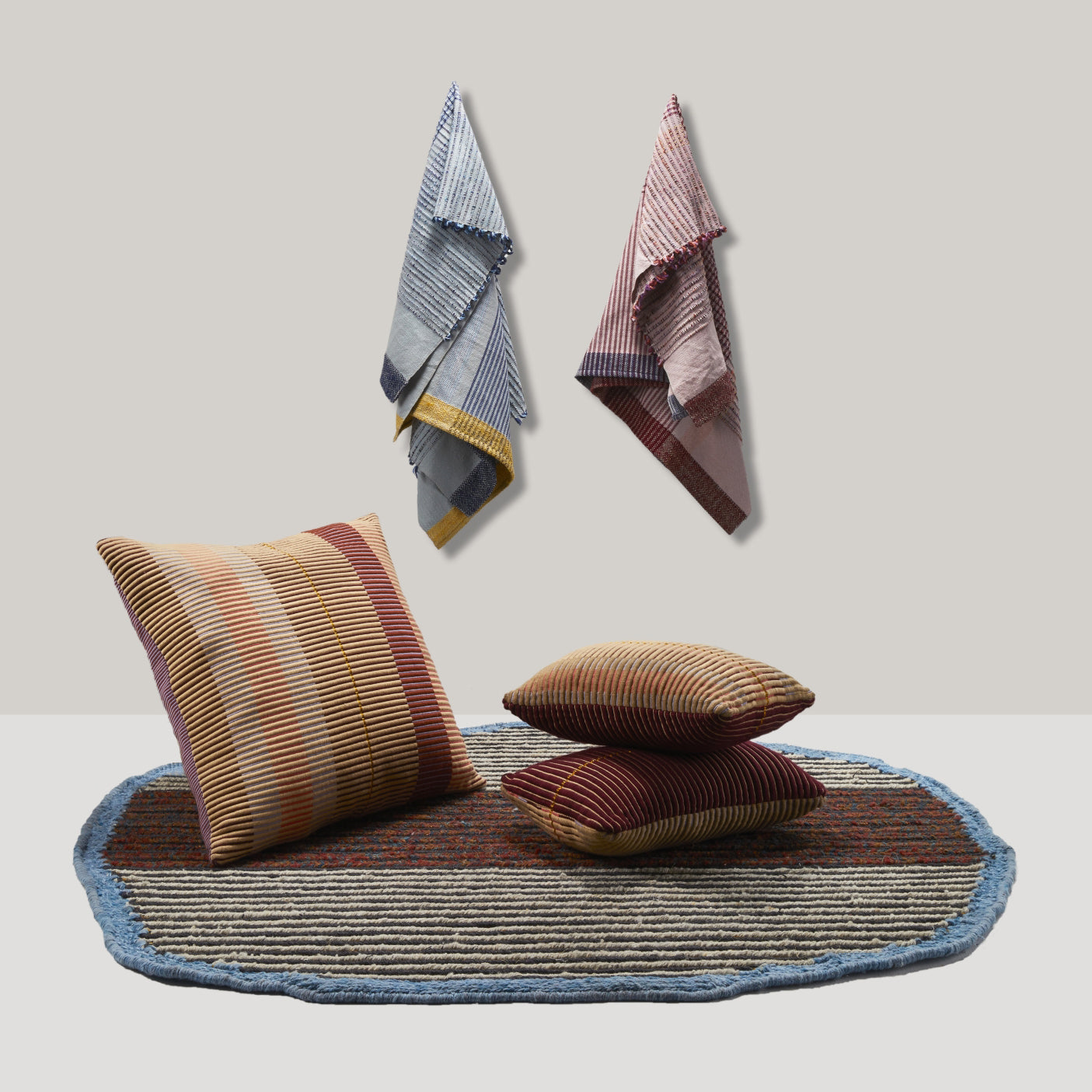 Three Ames Chumbes Cushions by Ames, each striped and hand-woven, sit on an oval rug with blue edges. Above them, three colorful striped cloths hang on a neutral, minimal wall.