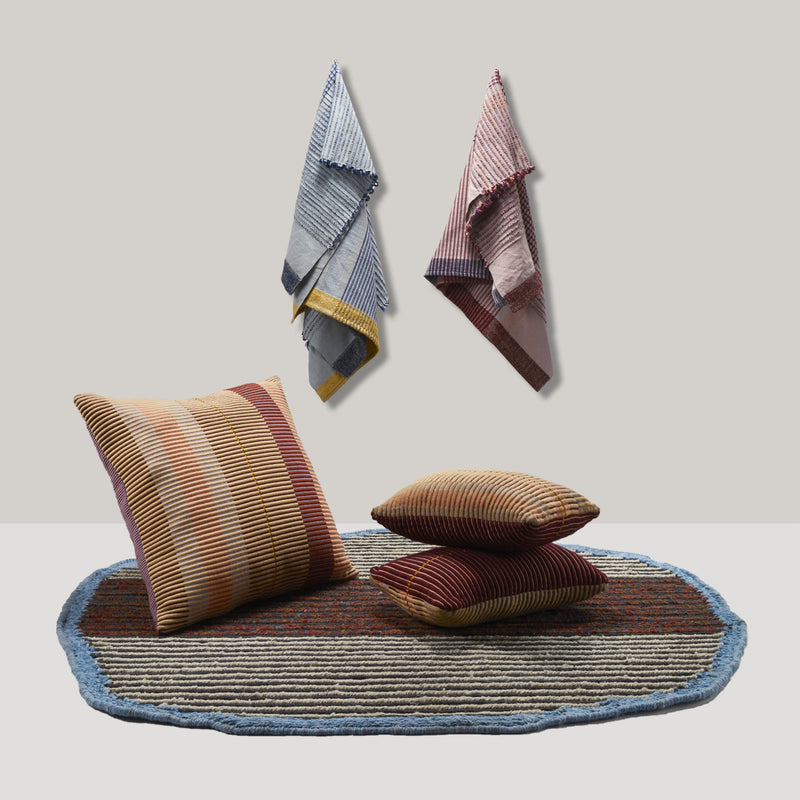 A round, textured rug with a blue border is adorned with three striped pillows and complemented by Ames Chumbes Throws hanging against a light wall in the background, showcasing shades of red, yellow, and brown.