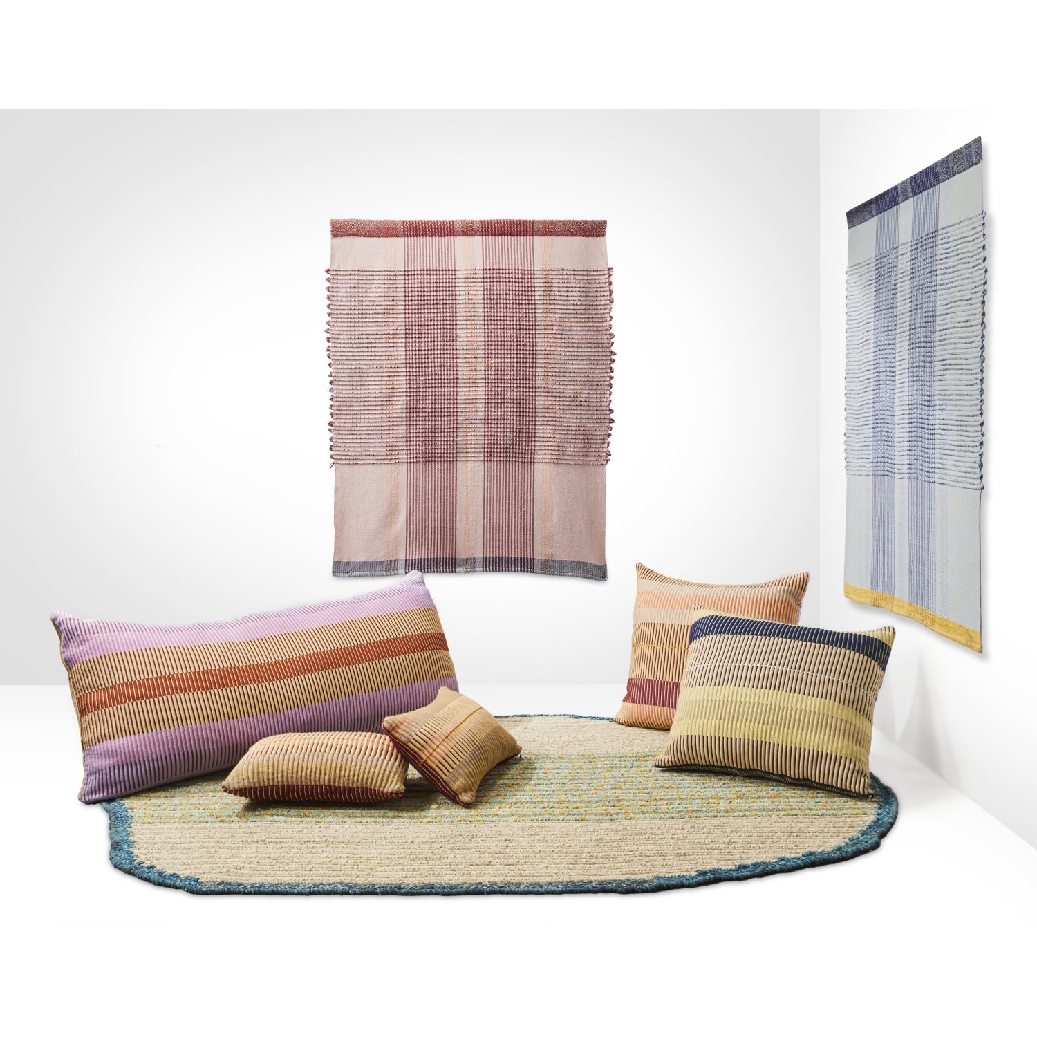 The image features a modern interior with two woven wall hangings and a circular rug. Five hand-woven Chumbes Layer cushions by Ames, in earthy tones and patterns, are placed around the rug, creating a cozy atmosphere.