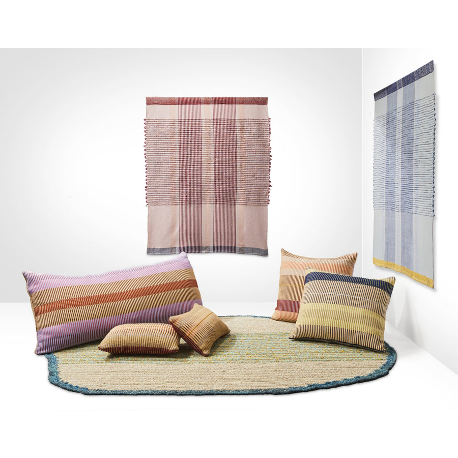 A modern room display features a round woven rug, five Ames Chumbes Cushions in earthy tones, and two striped textile wall hangings by Mae Engelgeer from Ames, all set against a plain white background.