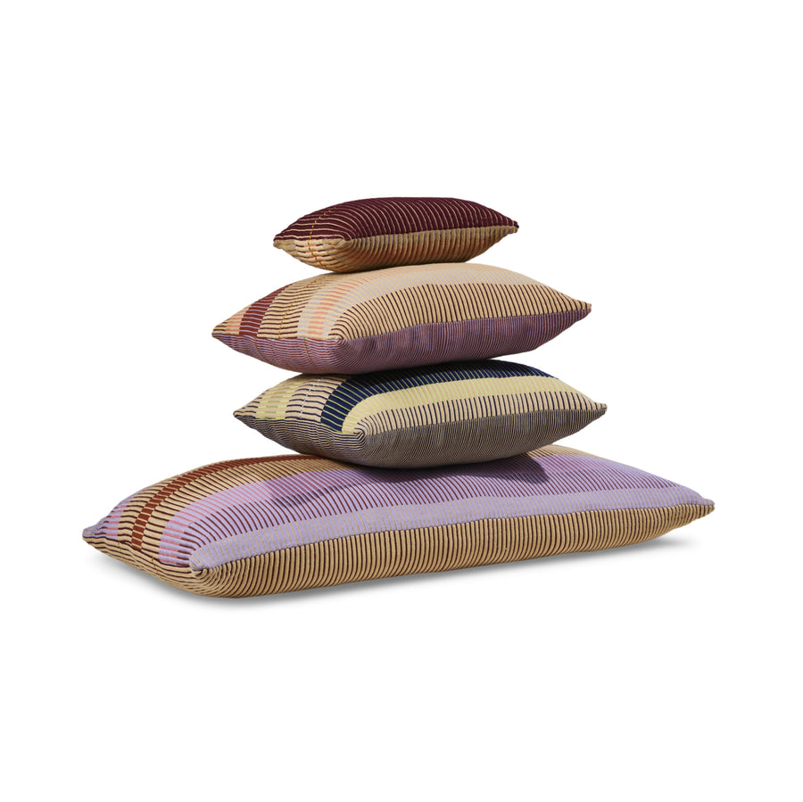 Four Ames Chumbes Layer pillows are stacked pyramidally against a white background. Each hand-woven cushion features a striped pattern in red, yellow, purple, and black, inspired by traditional textile symbols.