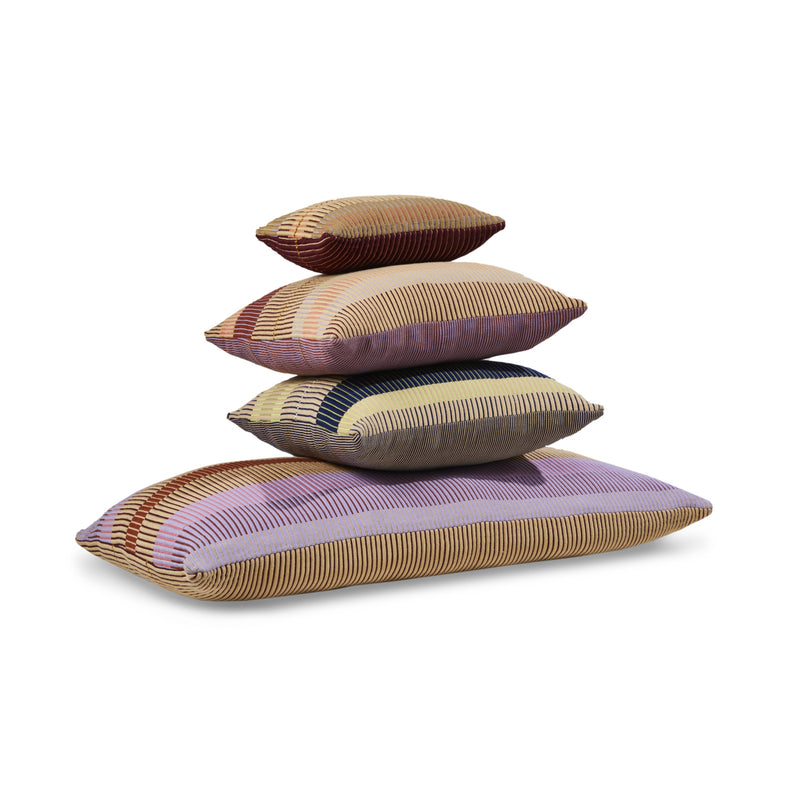 Four rectangular Ames Chumbes Cushions by Ames are stacked in different sizes, each with Mae Engelgeer’s signature stripes in beige, pink, purple, and blue on a white background.