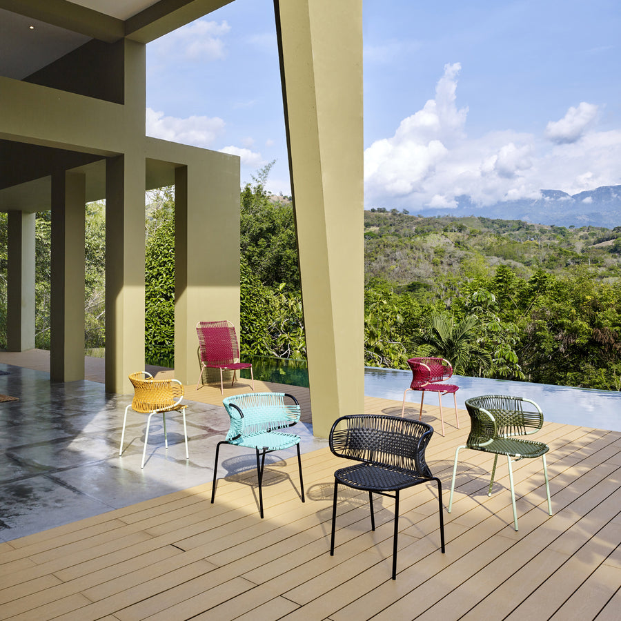 A modern outdoor seating area features the vibrant Cielo Armchairs by Ames, skillfully crafted with traditional weaving techniques. They sit on a wooden deck with lush greenery, a blue sky with clouds, distant mountains, and a swimming pool enhancing the contemporary architectural backdrop.