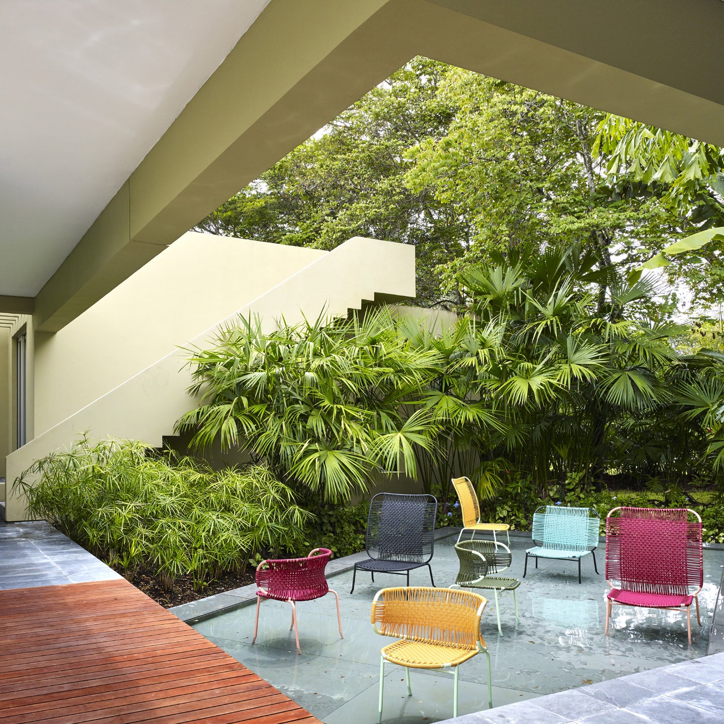 A modern outdoor area features a reflecting pool with Ames Cielo Armchairs, highlighting traditional weaving techniques. Surrounded by lush greenery, the architecture combines clean lines with wood and stone elements, creating a harmonious blend.