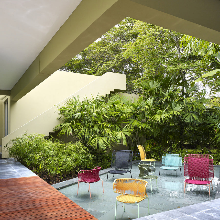 A modern outdoor area features a reflecting pool with Ames Cielo Armchairs, highlighting traditional weaving techniques. Surrounded by lush greenery, the architecture combines clean lines with wood and stone elements, creating a harmonious blend.