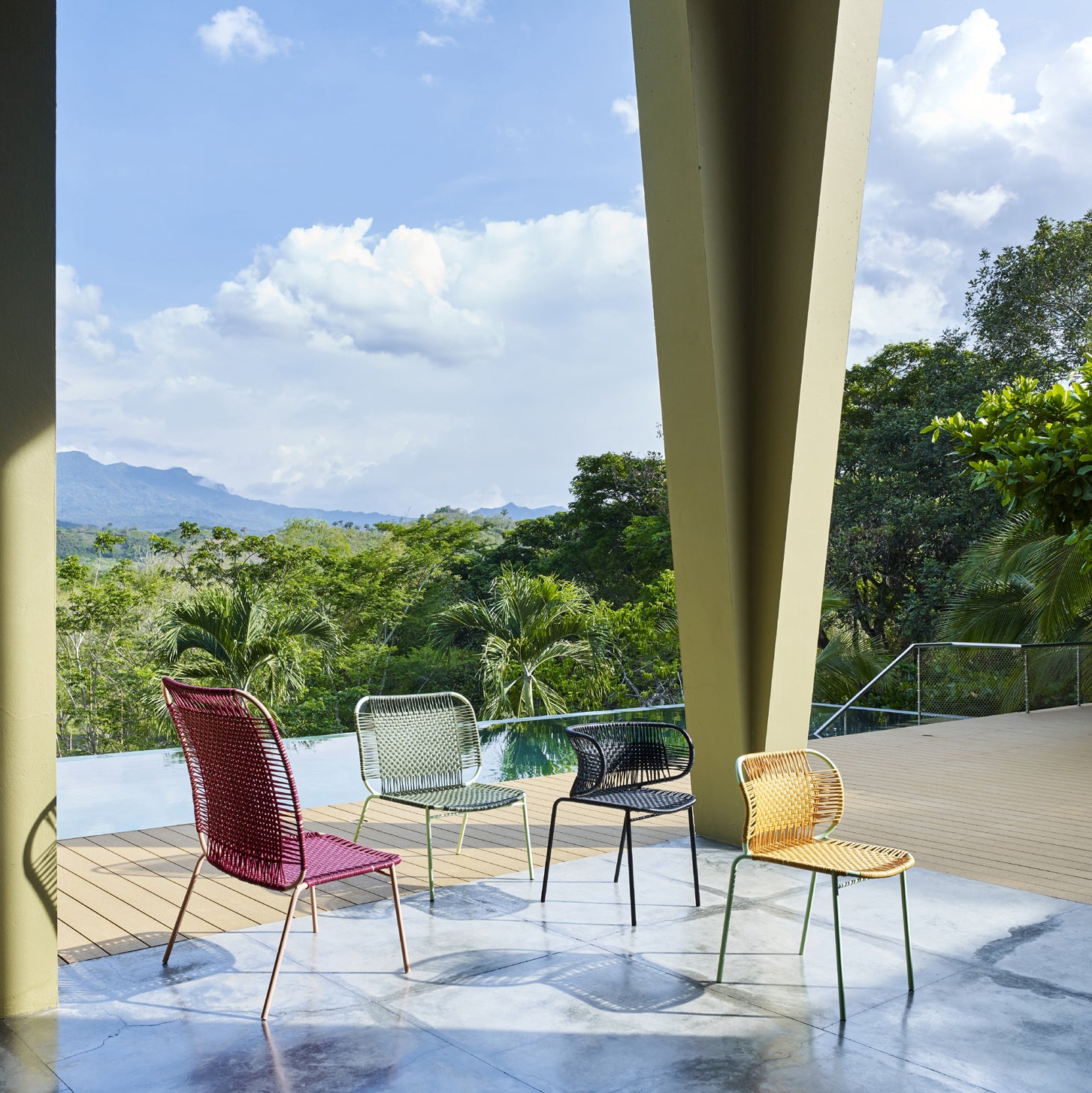 Four colorful Ames Cielo Lounge Chair Low by Ames are arranged outdoors on a patio with lush trees and mountains in the background under a partly cloudy sky. These stackable chairs are ideal for both indoor and outdoor use.