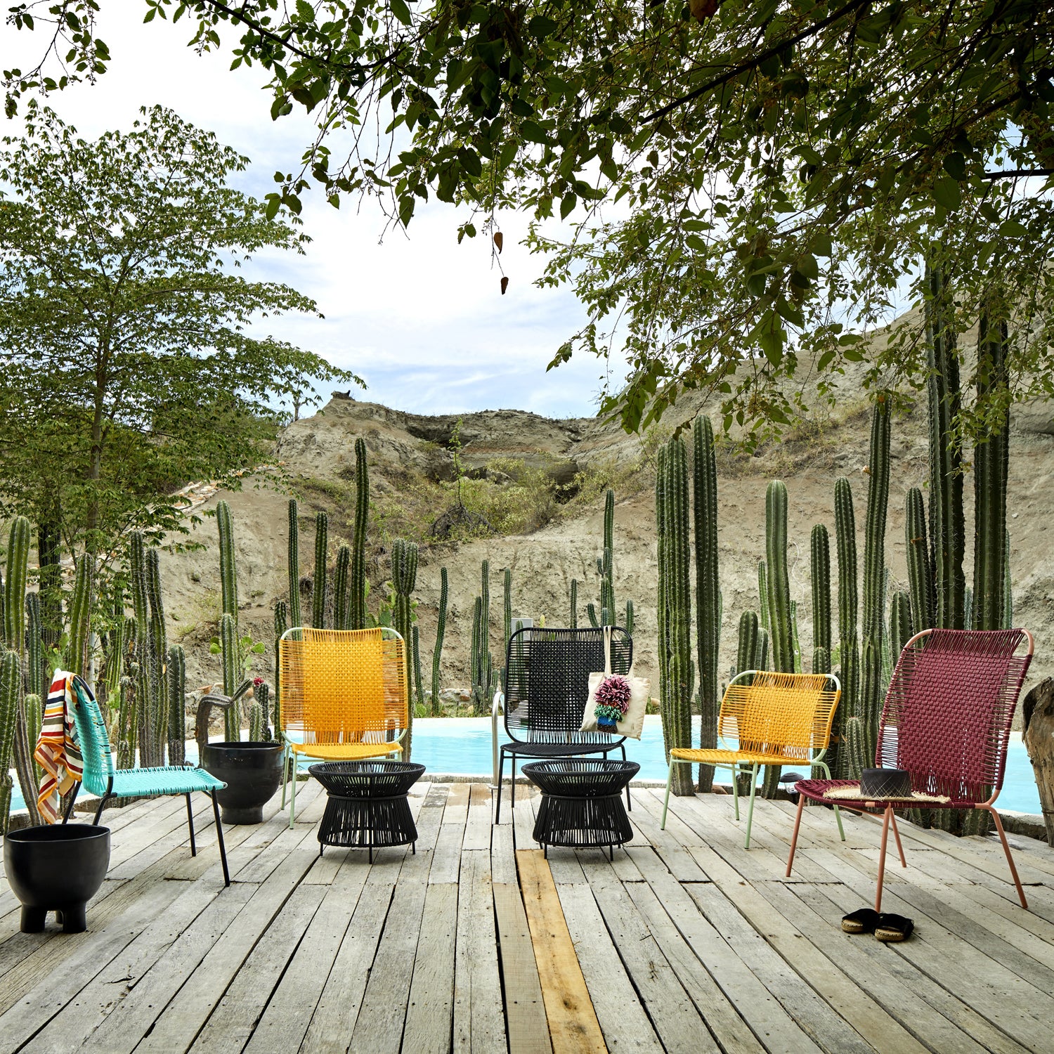 The Ames Cielo Lounge Chair High, featuring traditional weaving, is artfully placed on a wooden deck by the pool. Amidst tall cacti and desert hills, these vibrant chairs offer indoor-outdoor seating under the shade of tree branches.