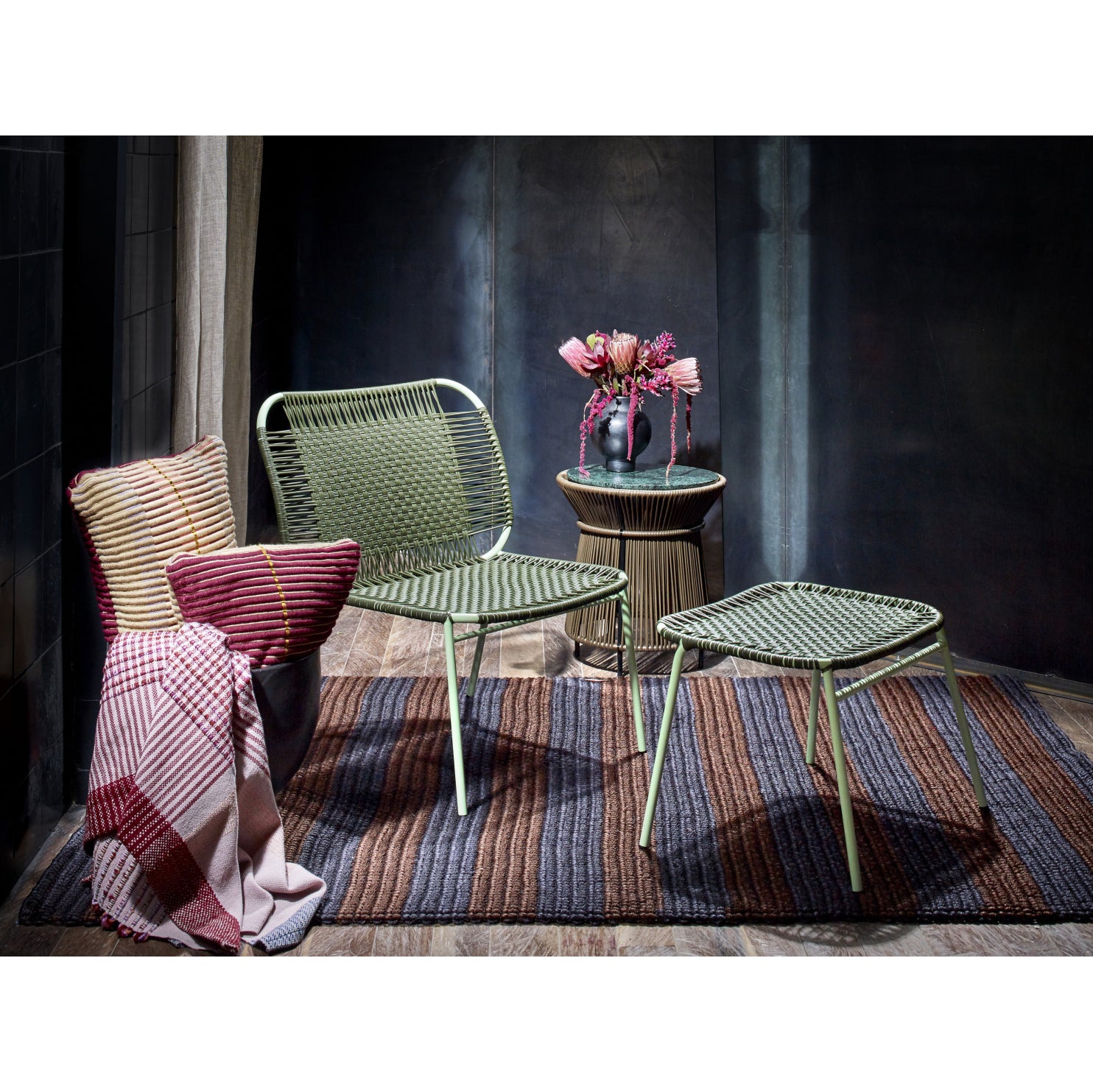 Two green woven chairs and matching Ames Cielo Ottomans by Ames, crafted with traditional weaving, rest on a striped rug. A small round table with pink flowers sits nearby; a plaid blanket and cushion are on the left chair in a cozy, dark room.