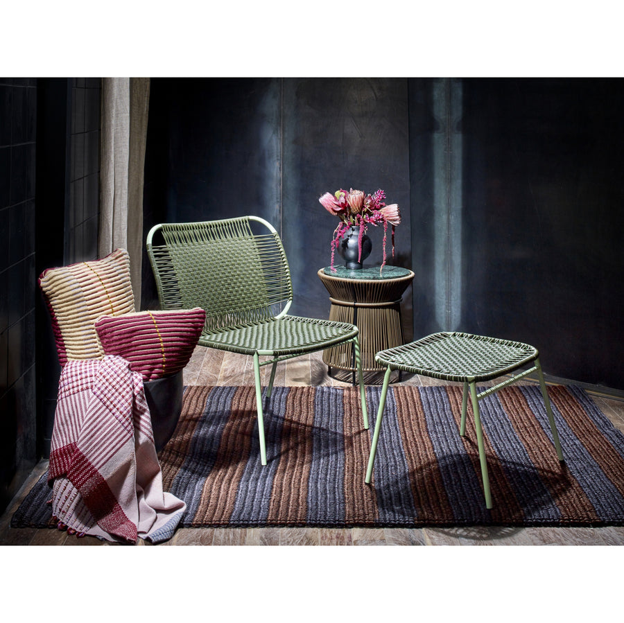 Two green woven chairs and matching Ames Cielo Ottomans by Ames, crafted with traditional weaving, rest on a striped rug. A small round table with pink flowers sits nearby; a plaid blanket and cushion are on the left chair in a cozy, dark room.
