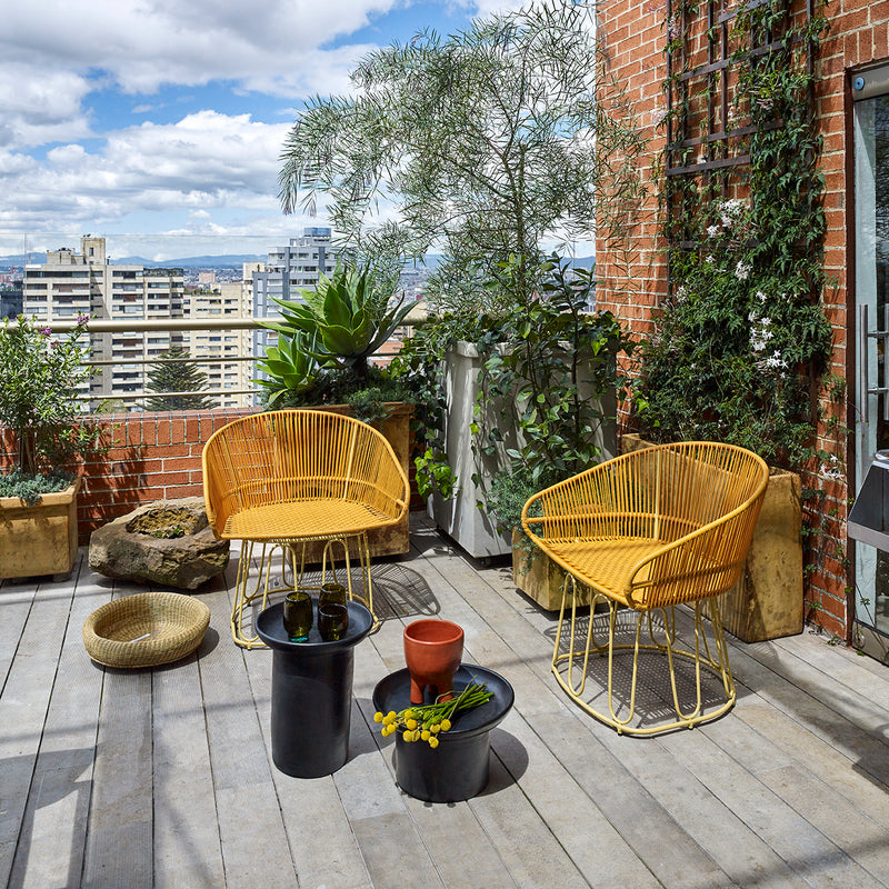 Rooftop patio with yellow Ames Circo Dining Chairs, small black tables, and vibrant green plants. A clay pot handcrafted by Colombian artisans displays yellow flowers on a table, with city buildings and a cloudy sky as the backdrop.