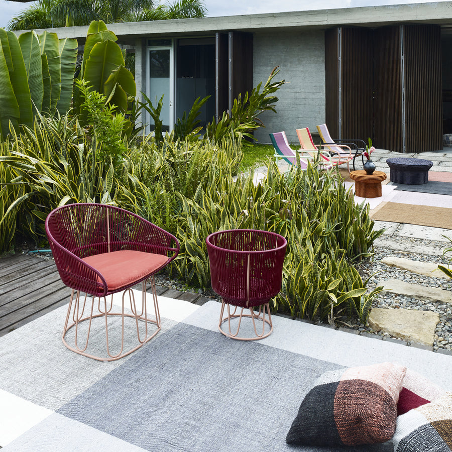 A modern patio showcases an Ames Circo Dining Chair and matching side table, set on a geometric-patterned rug. Crafted by Colombian artisans, lush green plants complement the scene with lounge chairs and a contemporary house in the distance.