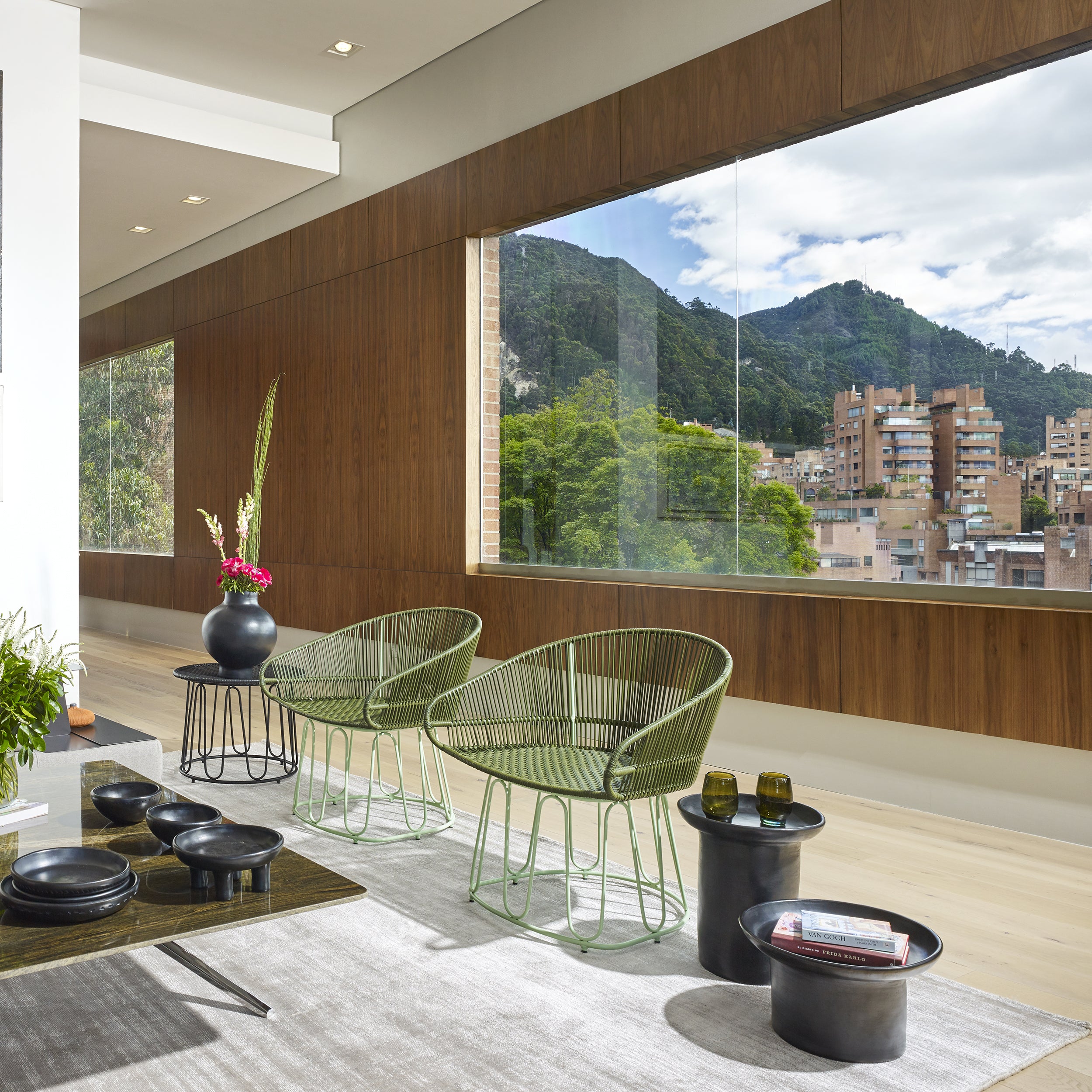 A modern room with large windows highlights a cityscape and green hills, featuring two green wire Circo Dining Chairs by Ames, a vase of flowers on a low table, all set on a gray rug. Wooden wall paneling showcases Colombian craftsmanship.