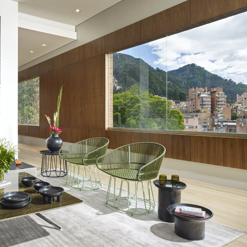 A modern room with large windows highlights a cityscape and green hills, featuring two green wire Circo Dining Chairs by Ames, a vase of flowers on a low table, all set on a gray rug. Wooden wall paneling showcases Colombian craftsmanship.