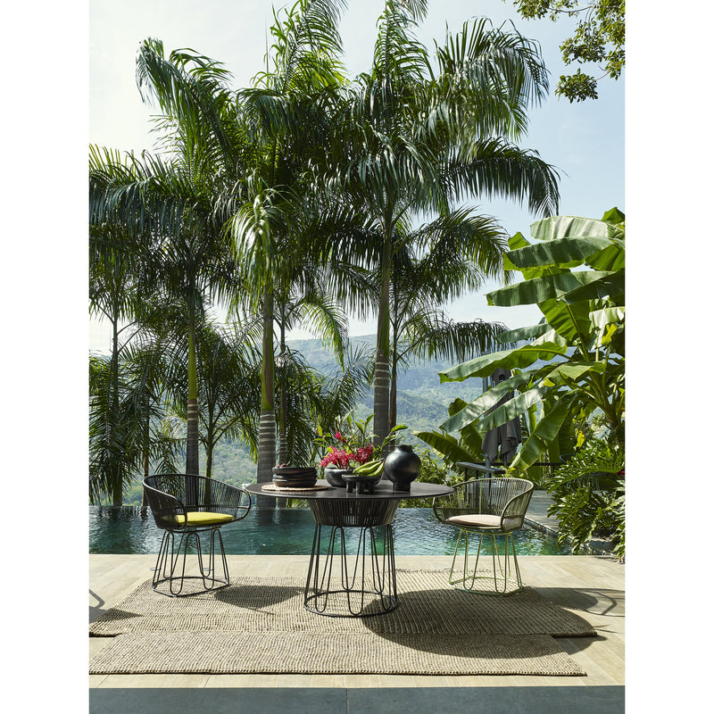 An outdoor dining area features a round table and four Ames Circo Dining Chairs on a woven rug. The handcrafted table displays a vase and fruit bowl, surrounded by lush greenery, tall palm trees, an infinity pool, and mountains in the background, showcasing Colombian artisans craftsmanship.