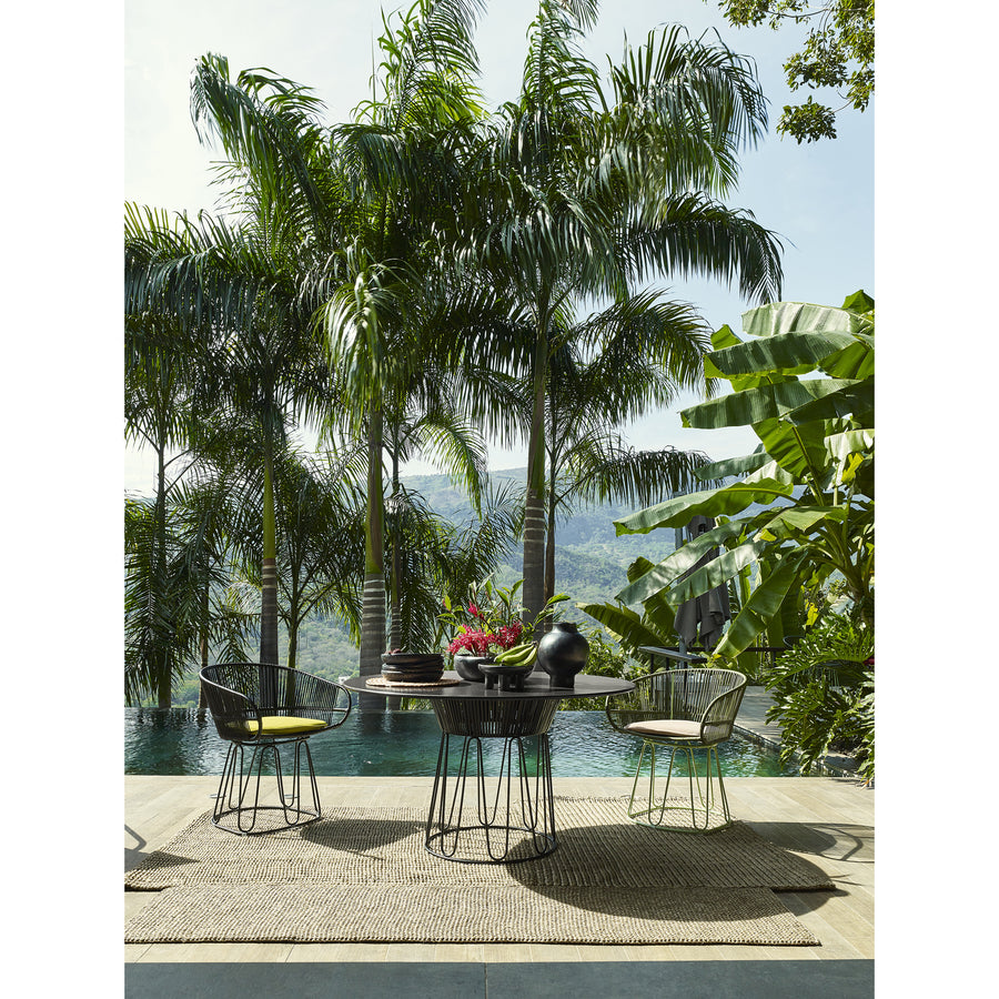 An outdoor dining area features a round table and four Ames Circo Dining Chairs on a woven rug. The handcrafted table displays a vase and fruit bowl, surrounded by lush greenery, tall palm trees, an infinity pool, and mountains in the background, showcasing Colombian artisans craftsmanship.
