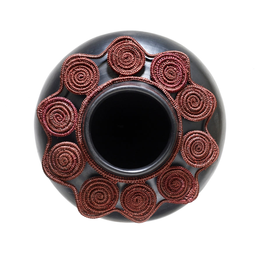 The Ames Coyar Vase 1 by Ames is a round black ceramic pot with a circular red coiled pattern resembling wickerwork around the opening, set against a white background.