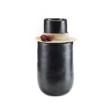 The Ames Coyar Vase 3 by Ames is a tall, smooth black ceramic vase with a wide base and narrow opening, adorned with a woven straw hat, two red ornaments, and wickerwork accents against a plain white background.