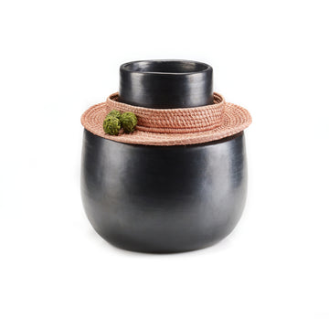 The Coyar Vase 4 by Ames is a matte black ceramic piece with a wide base and narrow top, accented by a Jipi palm wickerwork rattan collar. Two small green moss balls elegantly rest on the collar, adding natural contrast against the plain white backdrop.