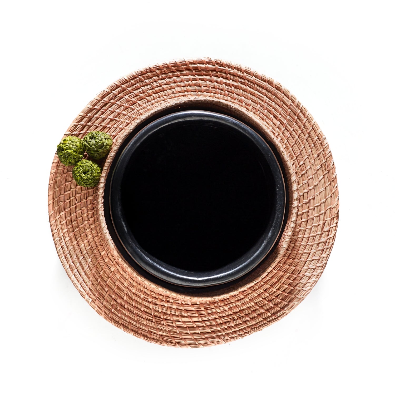 The Coyar Vase 4 by Ames, a black ceramic bowl, sits on a woven tan placemat. Two small green decorative items, possibly made from Jipi palm, are placed beside it. The image is shot from above against a white background.
