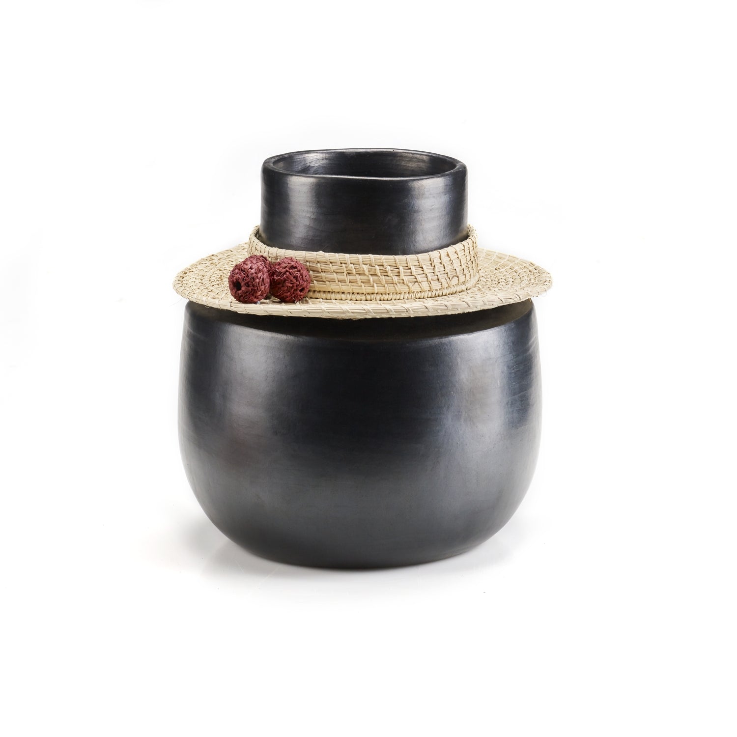 The Coyar Vase 4 by Ames is a matte black ceramic vase with a wide cylindrical body and narrow neck, adorned with a straw hat featuring Jipi palm wickerwork and red decorative flowers. It stands out vividly against a plain white background.