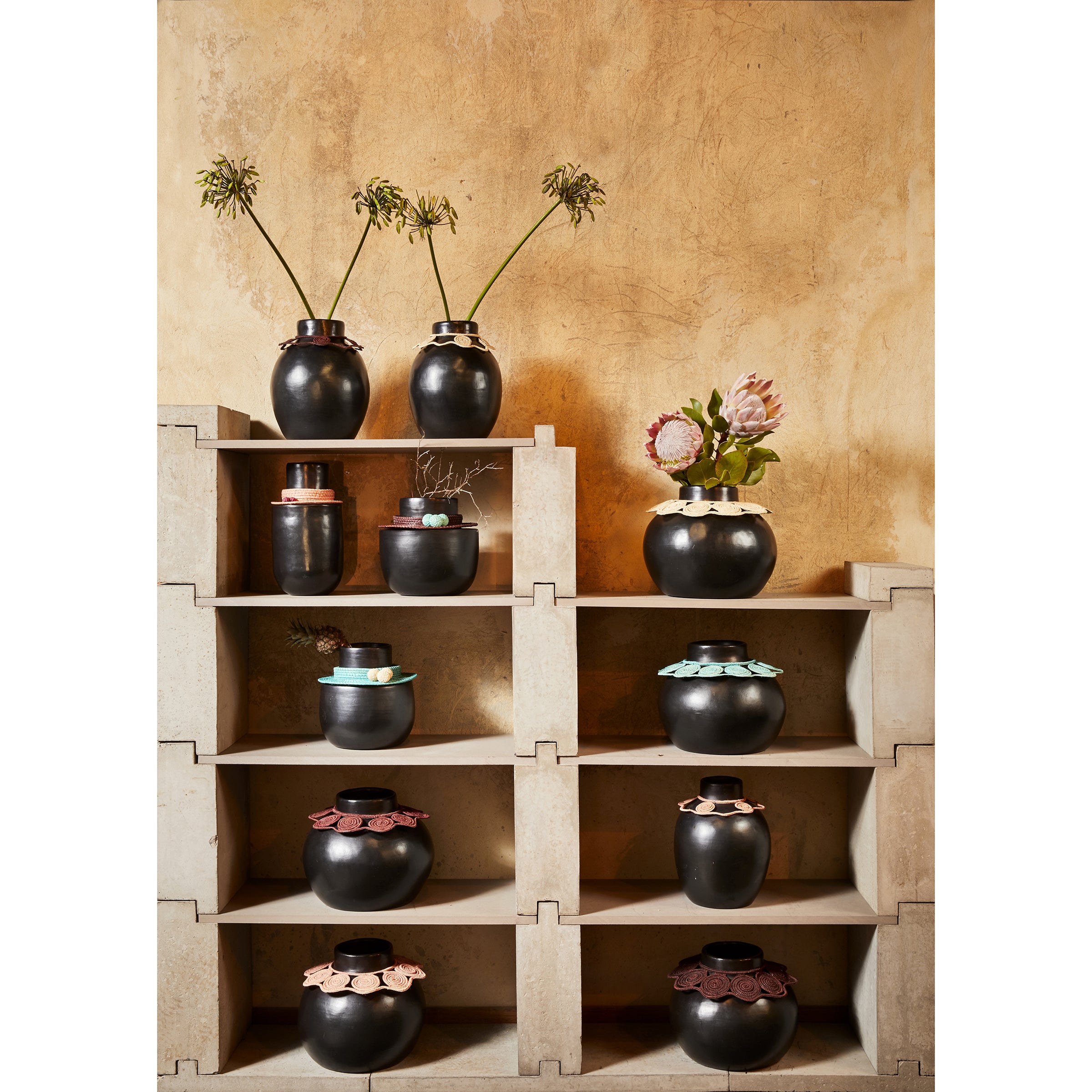 A display of Ames Coyar Vase 4 in black ceramic with colorful rims is stylishly arranged on a beige geometric shelf, featuring some vases filled with dried flowers and jipi palm accents against a textured, warm-toned wall.