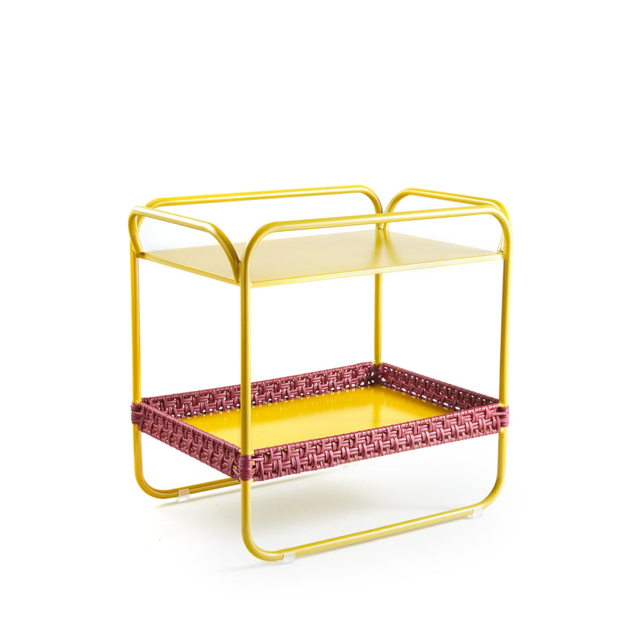 A small two-tier trolley with yellow shelves and a pink woven recycled plastic pattern around the bottom shelf, featuring rounded handlebars and minimalist design, perfectly complements Ames Dichas Sidetable.