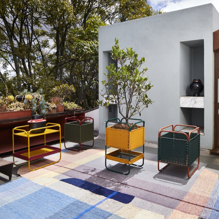 A modern outdoor patio features the Ames Dichas Natural Basket 1 by Ames, colorful woven cube planters, a geometric rug, potted succulents, a small tree, and a concrete fireplace set amid lush greenery under a partly cloudy sky.