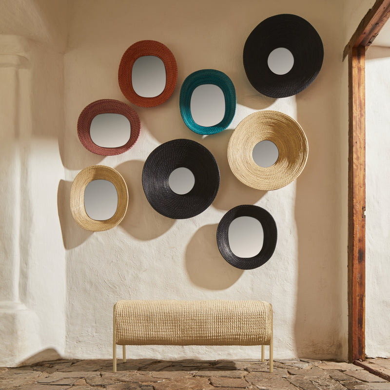 An Ames Mecato Bench by Ames rests beneath a wall adorned with oval mirrors framed in colorful iraca palm—black, tan, red, and teal—in a rustic room featuring textured white walls and a stone floor.