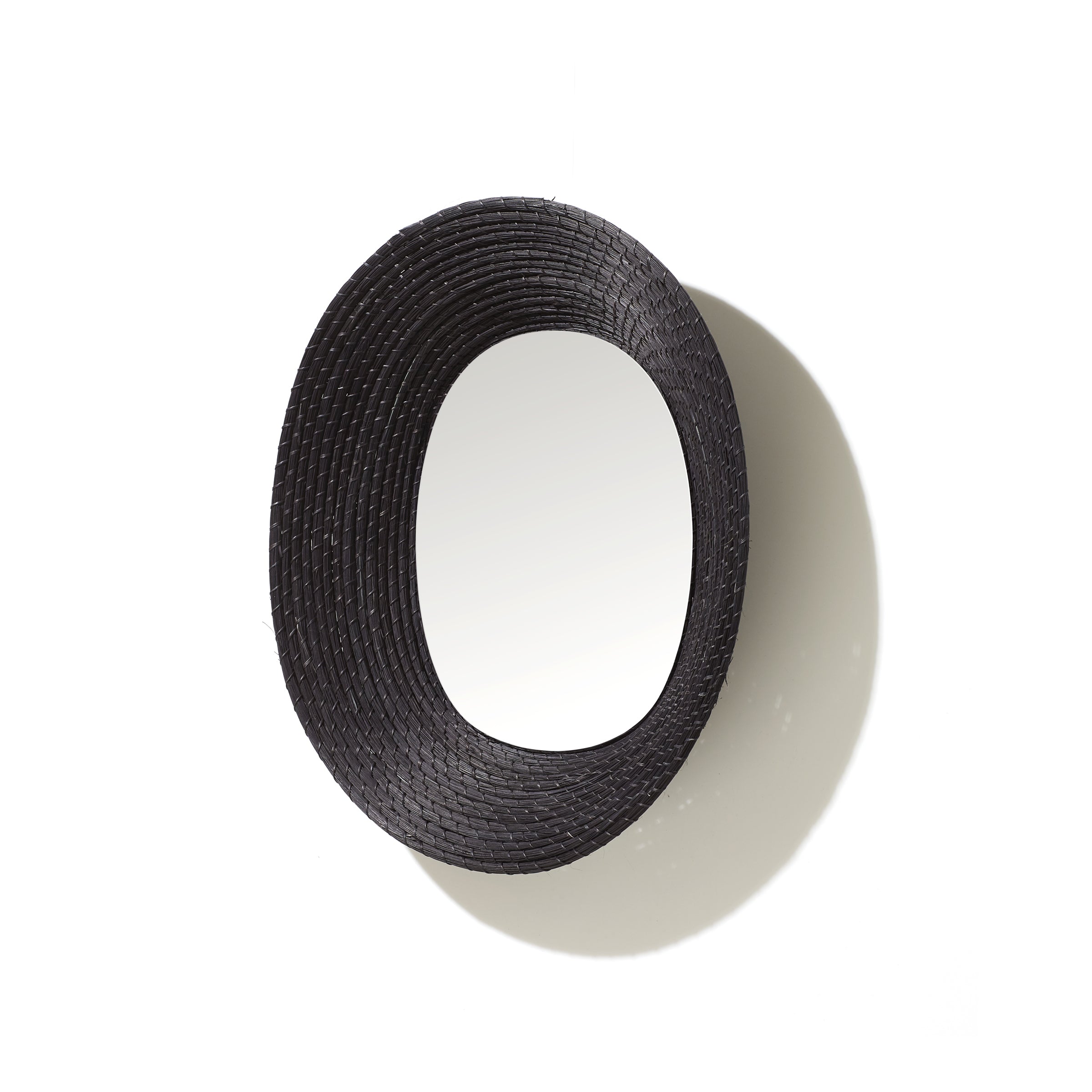 The Ames Killa Mirror by Ames features a black, oval-shaped wall design with a textured, handmade Iraca palm woven frame, casting a shadow on a white background.