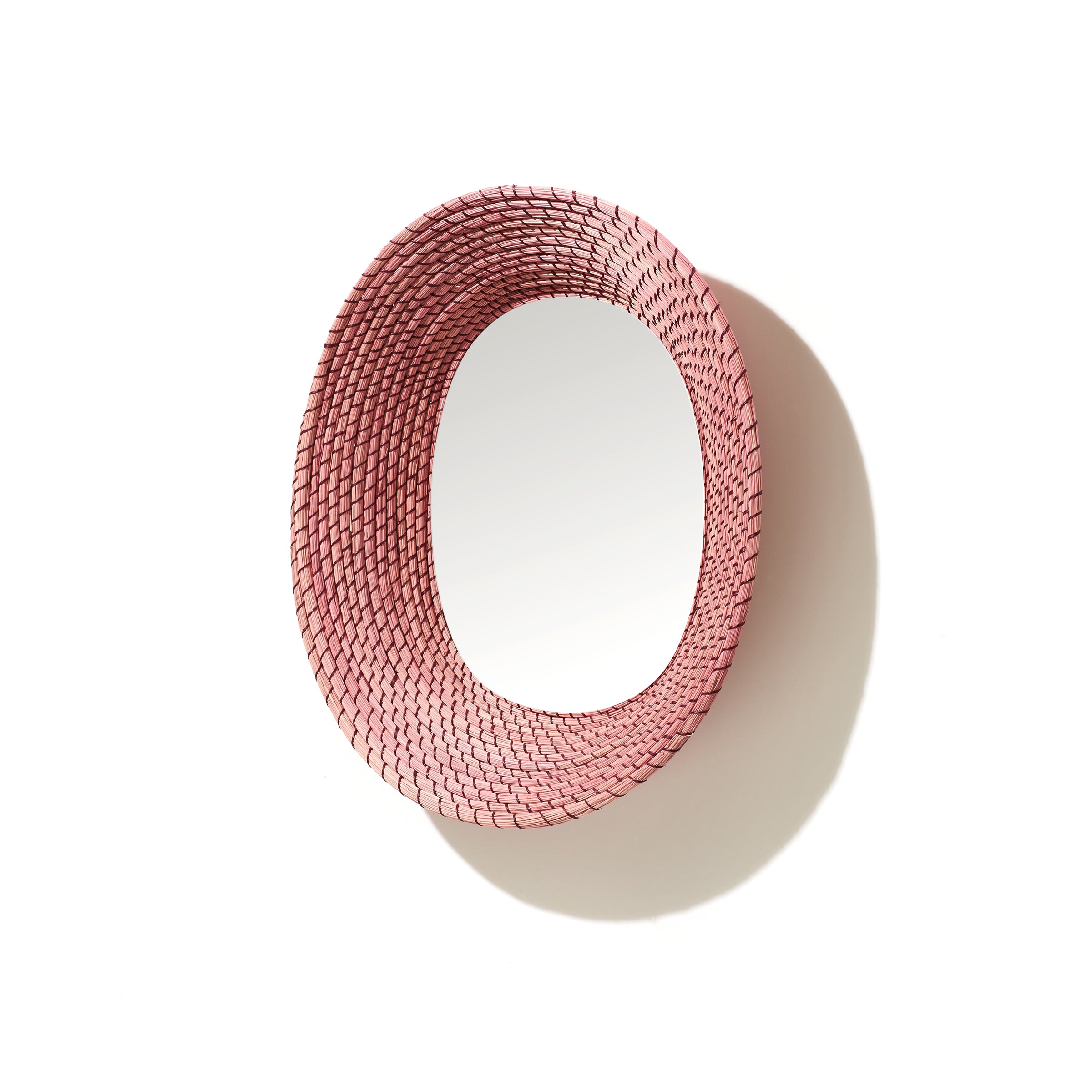 The Ames Killa Mirror by Ames features a handmade woven pink Iraca palm frame in a round design, casting a shadow on a white background.