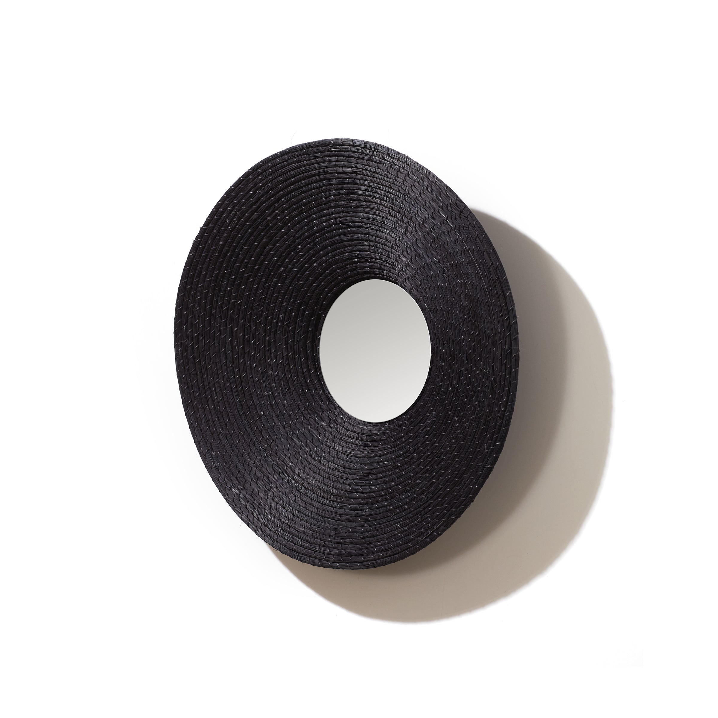 The Ames Killa Mirror by Ames features a round, handmade design with a wide, textured Iraca palm frame in black and a small central reflective surface, casting a soft shadow on a white background.