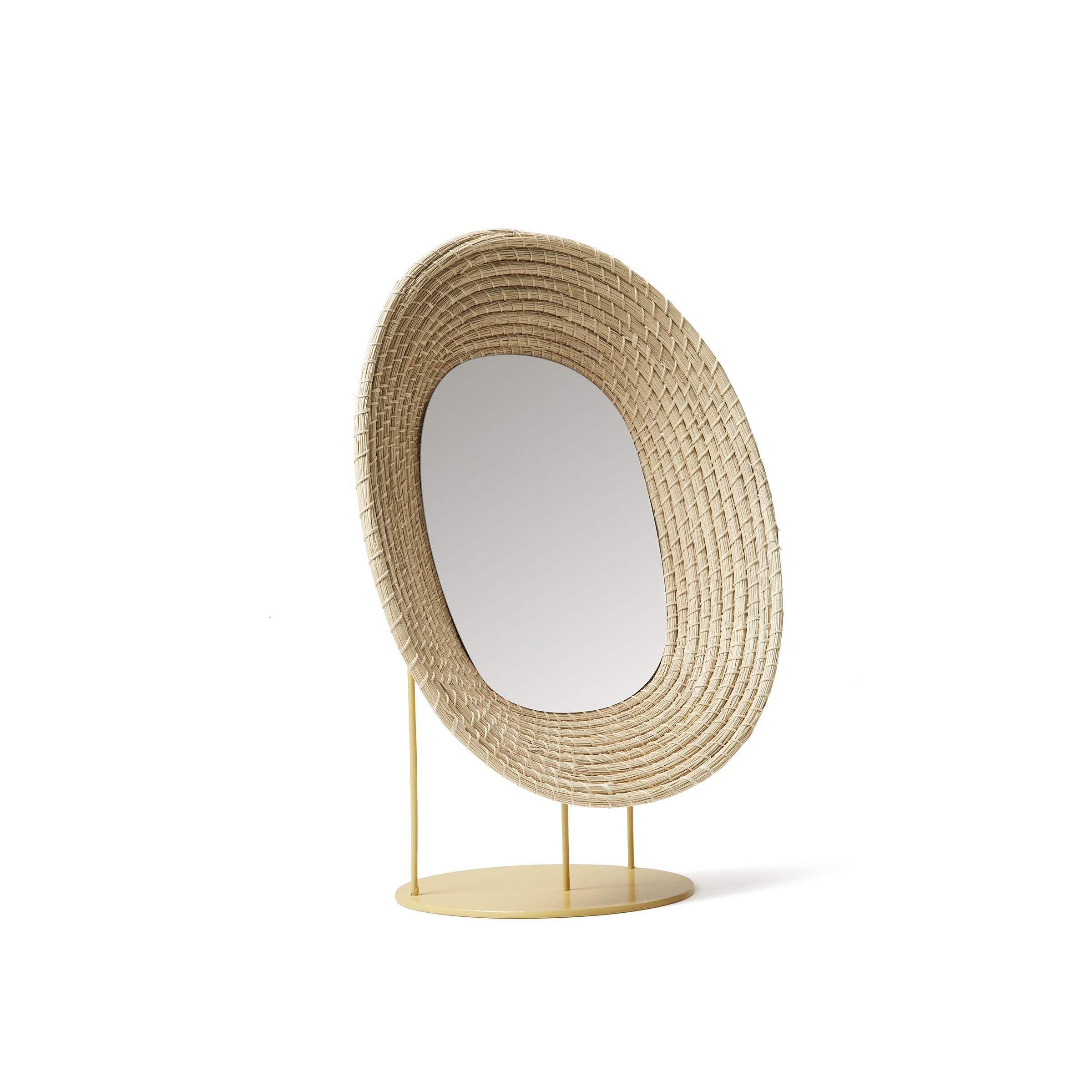 The Ames Killa Standing Mirror by Ames features an irregularly shaped woven iraca palm frame, set on a yellow circular metal base with slender supports.