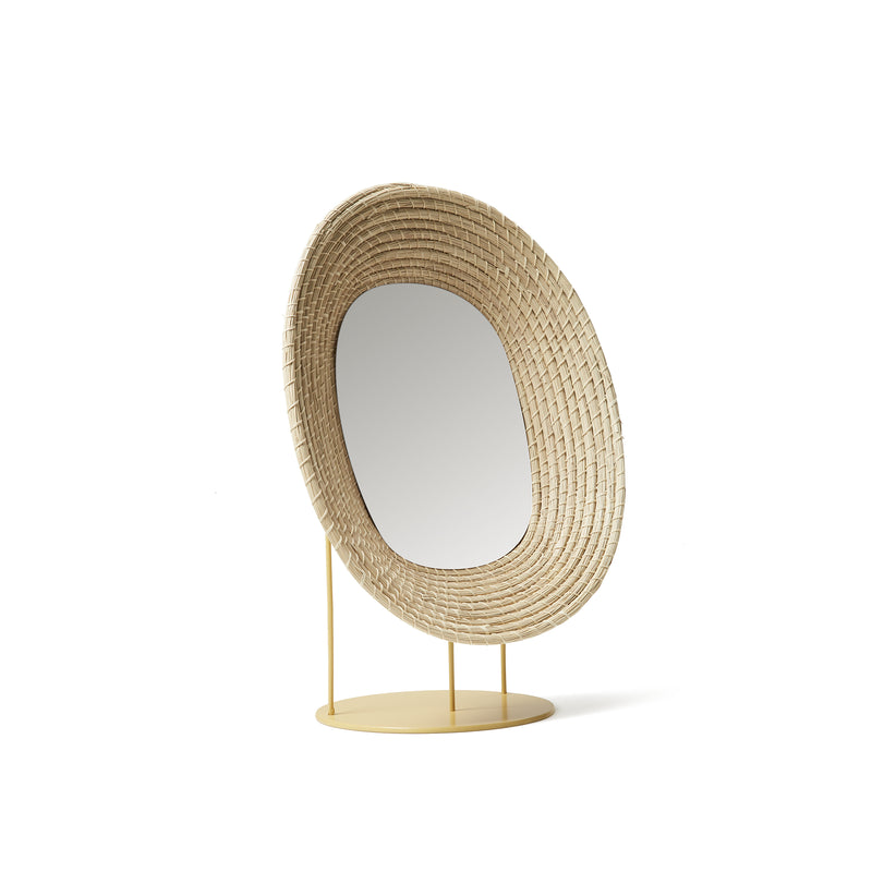 The Ames Killa Standing Mirror by Ames features an irregularly shaped woven iraca palm frame, set on a yellow circular metal base with slender supports.