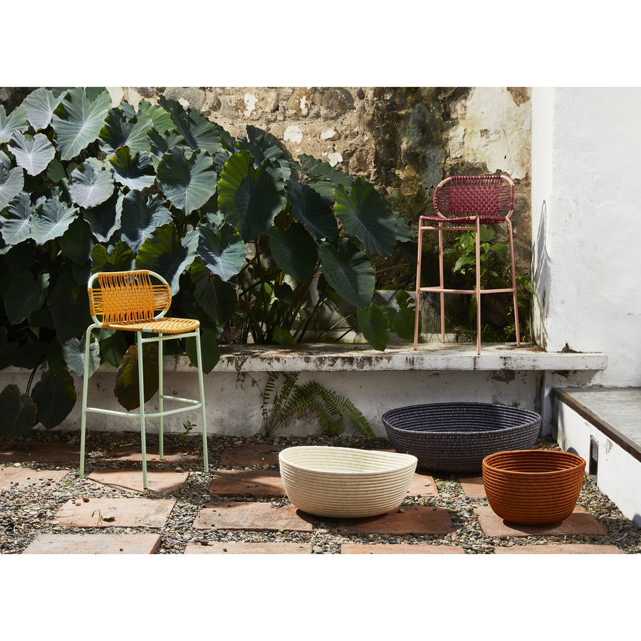 Four colorful woven chairs sit outdoors by a rustic wall and lush plants. In front, three round Ames La Che Baskets by Ames in white, black, and orange are displayed on a stone and gravel pathway.