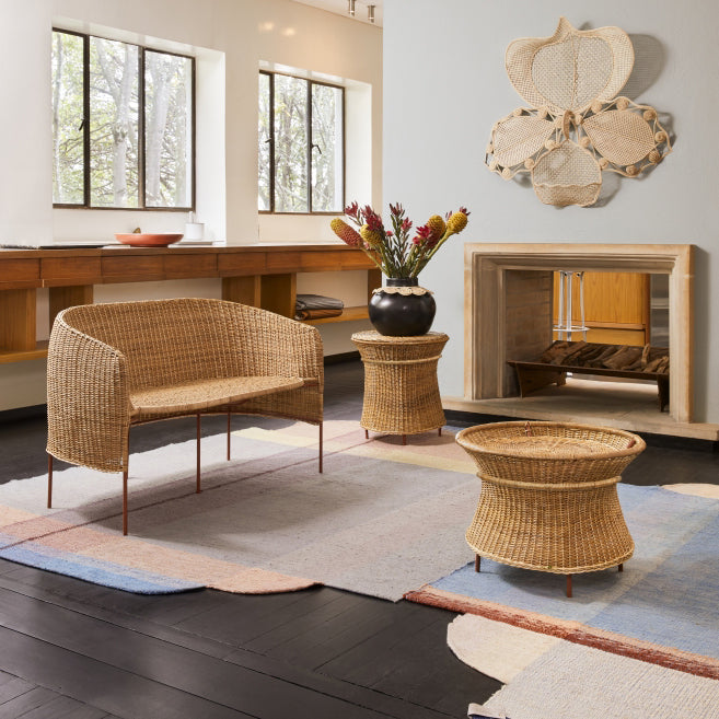 A cozy living room showcases wicker furniture, including a bench, side table, and coffee table on a multicolored rug. A floral arrangement with Coryanthes Macrantha adorns the side table. Large windows invite natural light, complemented by the Ames Macranta Wall Decor.