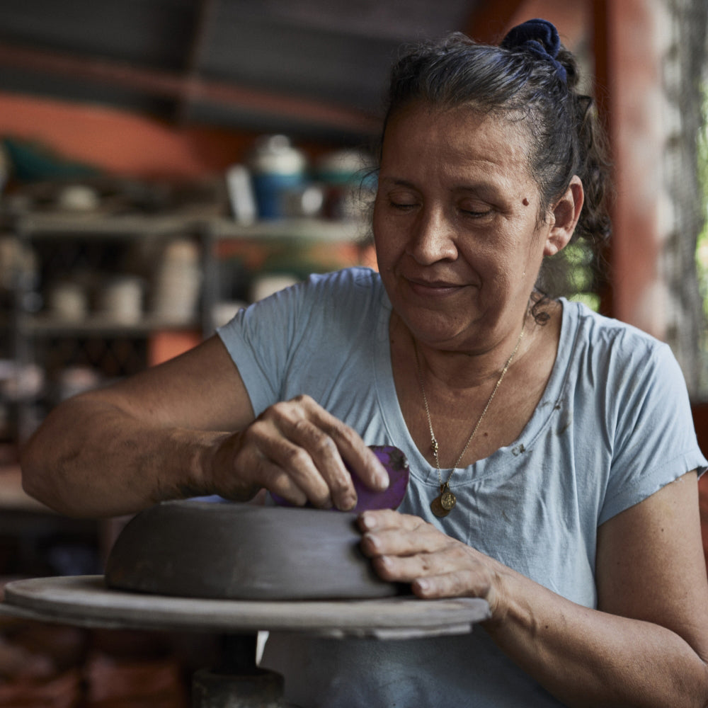 A woman in a light blue shirt is intently crafting Colombian pottery on a wheel, gently smoothing the black ceramic. Shelves behind her are lined with pieces from Ames Loza Ceramics, illustrating her artistic journey.